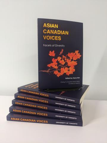 Image of the publication Asian Canadian Voices: Facets of Diversity book, shows a stack of books on a table with one book resting on top of the stack showcasing the front cover. The front cover says "Asian Canadian Voices Facets of Diversity" in gold font, on a dark background with orange maple tree branches in the middle.  